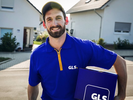 Livreur GLS souriant tenant un colis devant une maison, illustrant le service Pick&ShipService de collecte et livraison à l’adresse choisie.