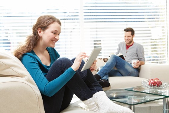 A man and woman sitting on a couch at home, the woman is looking at her tablet smilling.